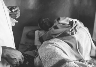 A Ugandan woman laying on a bed at a medical clinic while a doctor approaches